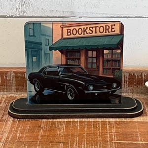 May include: A black classic car is parked in front of a bookstore, depicted on a wooden block. The bookstore has a green awning and the word "BOOKSTORE" is displayed on the sign. The block has a black, tiered base.