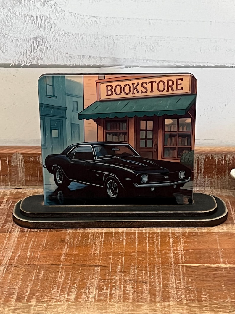 May include: A black classic car is parked in front of a bookstore, depicted on a wooden block. The bookstore has a green awning and the word "BOOKSTORE" is displayed on the sign. The block has a black, tiered base.