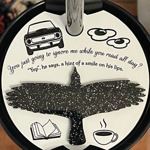 May include: A white tumbler lid with a black silhouette of a bird, a book, a cup of coffee, and two fried eggs. The text "You just going to ignore me while you read all day? "Yep", he says, a hint of a smile on his lips." is printed on the lid.