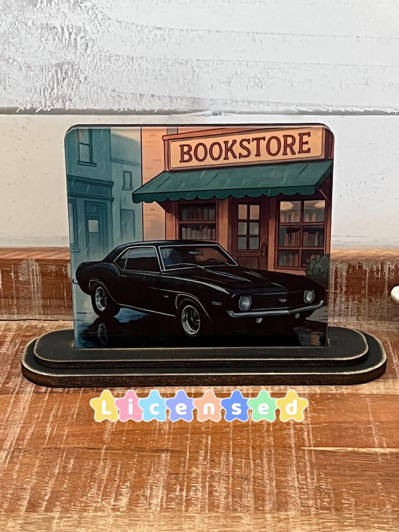 May include: A black classic car is featured in a bookstore scene. The image is on a wooden base. The word "BOOKSTORE" is visible on the sign above the store. The car is parked in front of the bookstore.