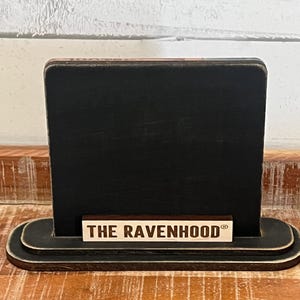 May include: A black wooden sign with a rectangular display area and a rounded base. The sign features the text "THE RAVENHOOD" in a light-colored font on a small rectangular piece of wood.