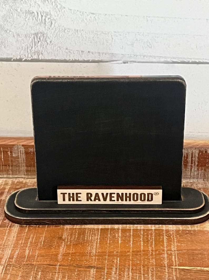 May include: A black wooden sign with a rectangular display area and a rounded base. The sign features the text "THE RAVENHOOD" in a light-colored font on a small rectangular piece of wood.