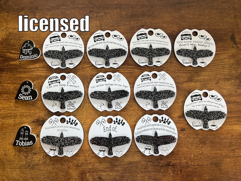 May include: Twelve black and white heart-shaped keychains with different sayings and images. The keychains feature a black bird with glitter, a car, a cup of coffee, a book, a wrench, a sun, a cloud, a chess piece, and a glass of wine. The keychains are on a wooden surface.