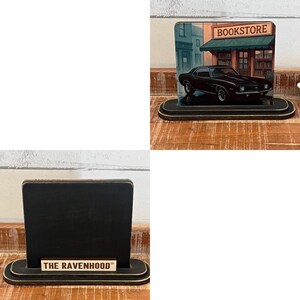 May include: Two wooden display stands. One features a black car parked in front of a bookstore. The other has a black chalkboard with the text "THE RAVENHOOD" on the base. Both stands have a black and gold color scheme.