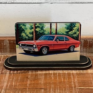 May include: A red classic car image on a wooden block. The car is a two-door coupe with chrome accents, set against a backdrop of trees. The block sits on a black, oval-shaped base, showcasing a vintage automotive theme.
