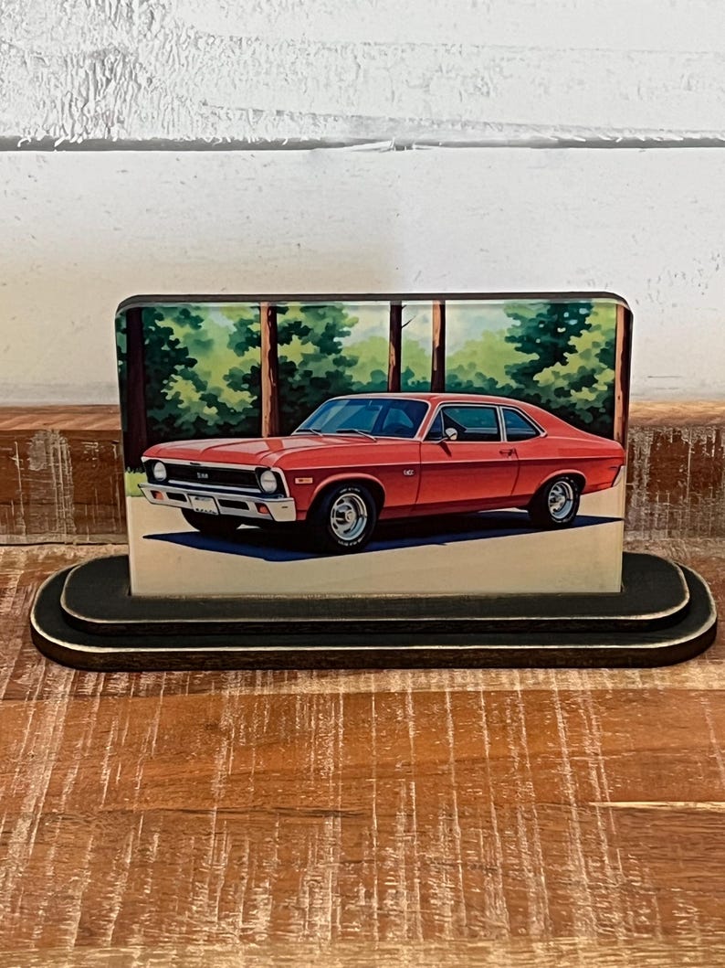 May include: A red classic car image on a wooden block. The car is a two-door coupe with chrome accents, set against a backdrop of trees. The block sits on a black, oval-shaped base, showcasing a vintage automotive theme.