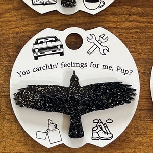 May include: A black glitter crow with outstretched wings on a white card with the text "You catchin' feelings for me, Pup?" and various black and white icons including a car, wrench, lighter, and hiking boot.