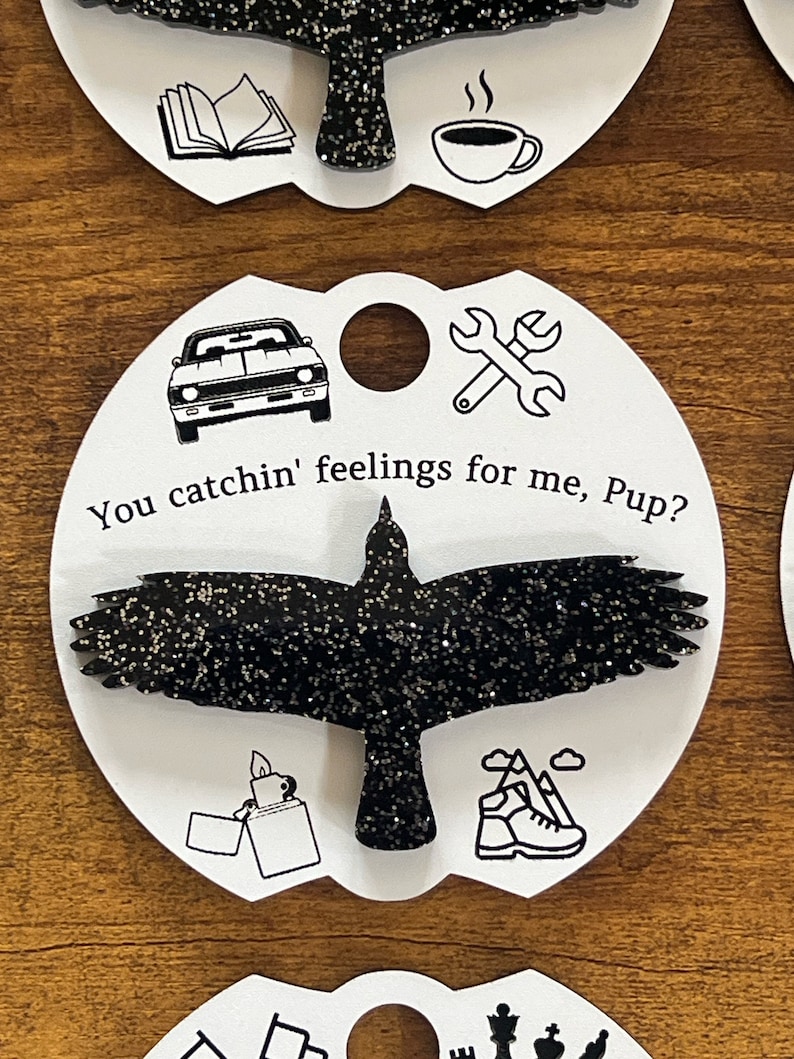 May include: A black glitter crow with outstretched wings on a white card with the text "You catchin' feelings for me, Pup?" and various black and white icons including a car, wrench, lighter, and hiking boot.