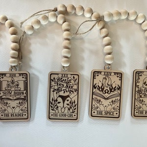 May include: Four wooden tarot card ornaments with white beads strung on twine. The cards feature illustrations and text, including "Slow Burn", "Yes Sir", "The Spice", and "Dark Romance".