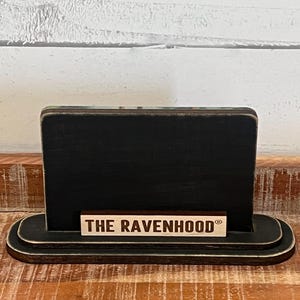 May include: A black wooden sign with a rounded base and a rectangular top. The sign has a small wooden plaque at the bottom that reads "THE RAVENHOOD" in capital letters. The sign is on a wooden surface.