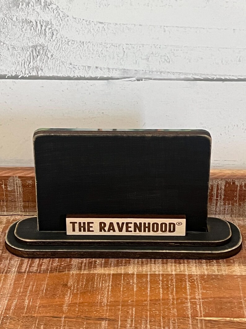 May include: A black wooden sign with a rounded base and a rectangular top. The sign has a small wooden plaque at the bottom that reads "THE RAVENHOOD" in capital letters. The sign is on a wooden surface.