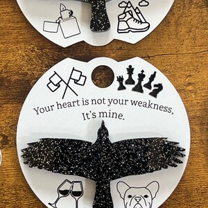 May include: Black glitter raven with outstretched wings on a white circle with the text "Your heart is not your weakness, It's mine." and various black and white icons around the raven.