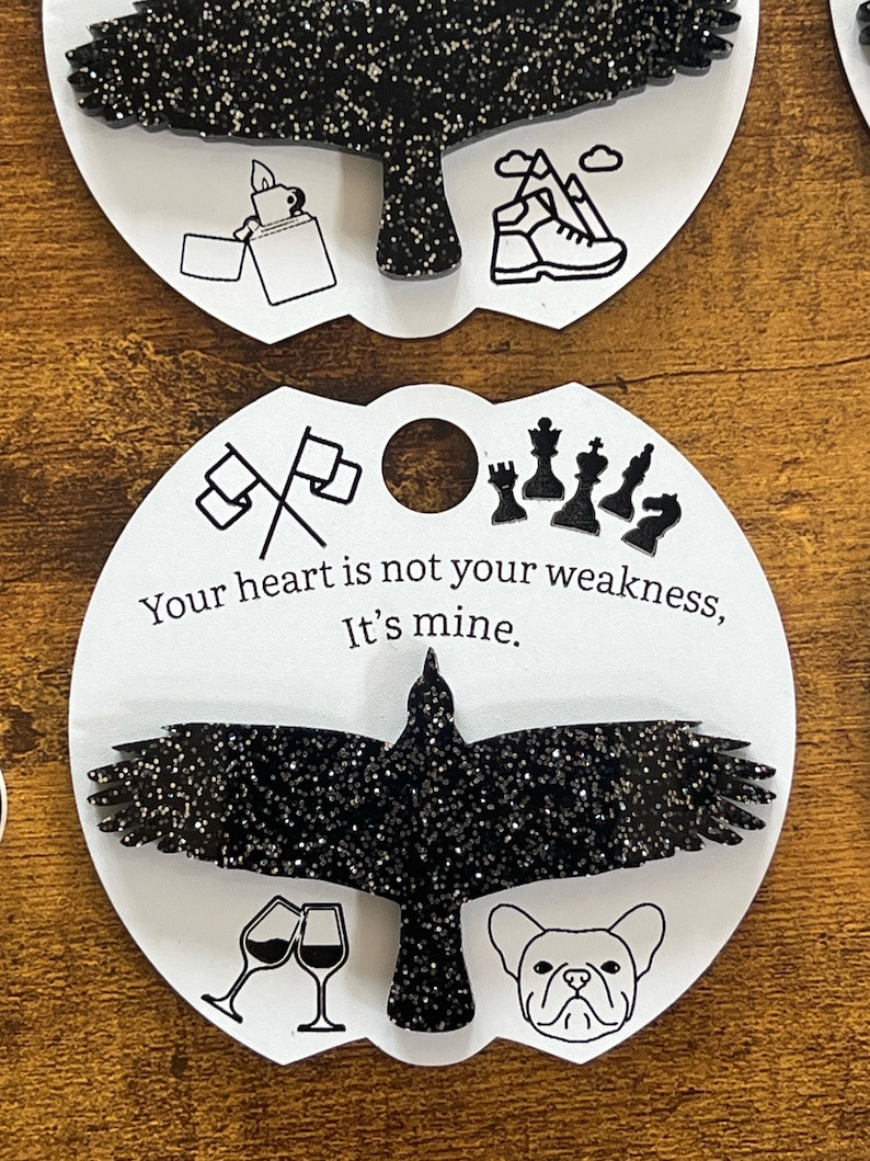 May include: Black glitter raven with outstretched wings on a white circle with the text "Your heart is not your weakness, It's mine." and various black and white icons around the raven.