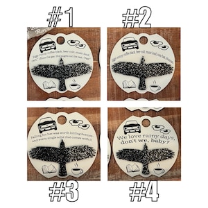 May include: Four white ceramic ornaments with black and white illustrations of a car, a raven, a book, and a coffee cup. Each ornament has a different quote printed on it. The quotes are: "Eggs-runny, coffee-black, beer-cold, music-loud. cars, "I floor the gas, she laughs out the rest. "Fast" ", "Eggs-runny, coffee-black, beer-cold, music-loud, cars-fast, Woman", "Falling for her was worth hitting bottom- and every single ache that comes with it.", and "We love rainy days, don't we, baby?"