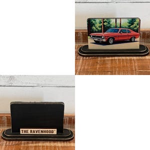 May include: A black wooden desk organizer with a rounded base and a rectangular back. The organizer features a printed image of a red car. The text "THE RAVENHOOD" is printed on the base.