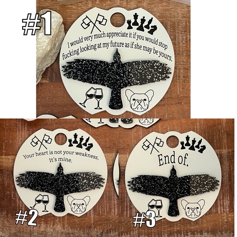 May include: Three white circular ornaments with black glitter accents. Each ornament features a black silhouette of a bird with a French bulldog, a pair of wine glasses, and a set of chess pieces. The text on the ornaments reads: "I would very much appreciate it if you would stop fucking looking at my future as if she may be yours.", "Your heart is not your weakness. It's mine.", and "End of."