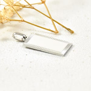 Puede incluir: Un colgante rectangular de plata con un borde texturizado y un bucle en la parte superior para sujetar una cadena.