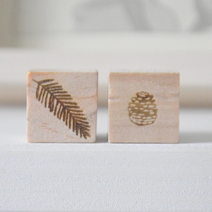 Puede incluir: Dos pequeños bloques de madera cuadrados. Uno presenta un diseño de helecho marrón, y el otro un diseño de piña marrón. Los bloques son de color claro y parecen hechos a mano, perfectos para manualidades o fines decorativos.