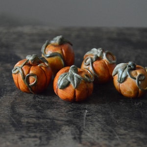 Op de afbeelding: Vijf kleine, ronde, oranje keramische pompoenen met getextureerde oppervlakken en gebeeldhouwde groene stelen en ranken. Elke pompoen heeft verticale ribbels en een handbeschilderde afwerking, gerangschikt in een groepje.