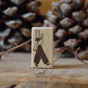 Puede incluir: Un pequeño bloque de madera rectangular con una ilustración tallada de una persona que lleva leña. La figura es de color marrón oscuro sobre la veta de madera clara. El fondo incluye dos piñas grandes y una superficie de madera.