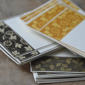 May include: A stack of decorative cards with floral patterns. The cards feature a white base with decorative borders in gold, brown, and yellow. The floral designs include leaves and other botanical elements. The cards are stacked on a wooden surface.