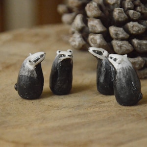 Op de afbeelding: Kleine keramische dassenbeeldjes met zwart-witte markeringen. De dassenbeeldjes zijn gerangschikt op een houten oppervlak, met een dennenappel op de achtergrond. De beeldjes zijn ongeveer 4 cm hoog.