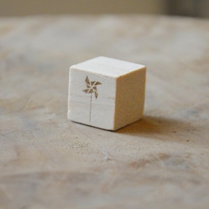 Puede incluir: Un pequeño bloque de madera cuadrado con un diseño de molinillo grabado en un lado. El molinillo es marrón y el bloque es de color beige claro. El bloque está sobre una superficie marrón claro.