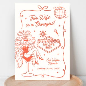 Op de afbeelding: Een crèmekleurige print met oranje illustraties en tekst. Het ontwerp toont een showgirl in een martiniglas, een discobal en de woorden "This Wife is a Showgirl!" en "Welcome Taylor's Bach" met locatie en datum.