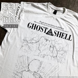 May include: White t-shirt featuring a black graphic design from "Ghost in the Shell." The design includes the title, a quote, and an illustration of a person. The Etsy shop name "EELS AND BRO" is also visible.