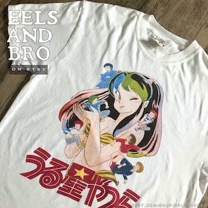 Puede incluir: Camiseta blanca con un gráfico de anime colorido. El diseño muestra un personaje femenino con cabello verde y azul, rodeado de figuras más pequeñas. Texto japonés en rojo. El texto "EELS AND BRO" está impreso en la esquina superior izquierda.