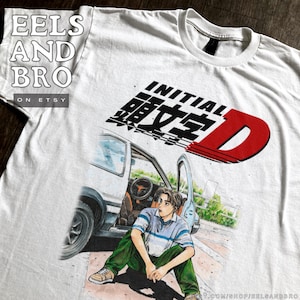Puede incluir: Camiseta blanca con un gráfico de un coche y una persona, con el texto "INITIAL D" en negro y rojo. La persona lleva una camisa a rayas azules y blancas y pantalones verdes. La camiseta tiene el texto "EELS AND BRO".