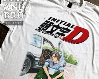 May include: White t-shirt featuring a graphic of a car and a person, with the text "INITIAL D" in black and red. The person is wearing a blue and white striped shirt and green pants. The shirt has the text "EELS AND BRO" on the top left.