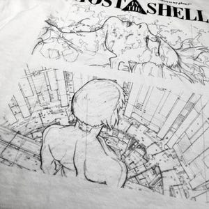 May include: A white t-shirt featuring a black ink-style graphic print. The design includes the text GHOST IN THE SHELL, a sketch of a face with flowing hair, and a rear-view illustration of a figure overlooking a detailed urban cityscape.