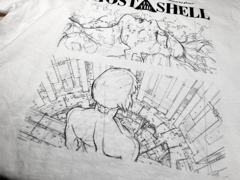May include: A white t-shirt featuring a black ink-style graphic print. The design includes the text GHOST IN THE SHELL, a sketch of a face with flowing hair, and a rear-view illustration of a figure overlooking a detailed urban cityscape.