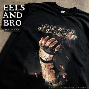 May include: Black t-shirt featuring the text "DEAD SPACE" in a glowing, distressed font. The graphic includes a detailed image of a robotic arm with blood splatter. The Etsy shop name "EELS AND BRO" is visible.