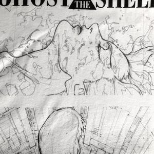 May include: A white t-shirt featuring a black ink-style illustration of a figure looking upward with flowing hair, surrounded by abstract geometric patterns. The text GHOST IN THE SHELL is printed in black capital letters at the top of the design.