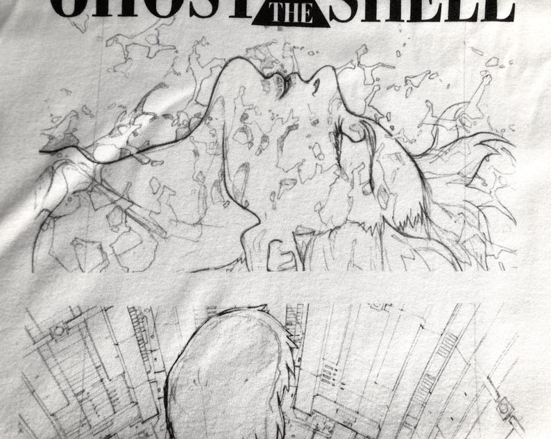 May include: A white t-shirt featuring a black ink-style illustration of a figure looking upward with flowing hair, surrounded by abstract geometric patterns. The text GHOST IN THE SHELL is printed in black capital letters at the top of the design.