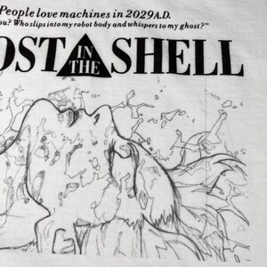 May include: A white t-shirt featuring a black line art illustration of a figure with flowing hair. The text reads: People love machines in 2029 A.D. Who slips into my robot body and whispers to my ghost? GHOST IN THE SHELL is printed in black.