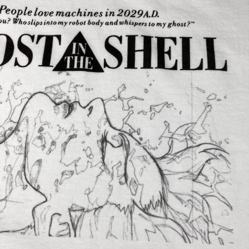 May include: A white t-shirt featuring a black line art illustration of a figure with flowing hair. The text reads: People love machines in 2029 A.D. Who slips into my robot body and whispers to my ghost? GHOST IN THE SHELL is printed in black.