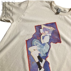 May include: Cream-colored t-shirt featuring a graphic of an anime character in a blue and white outfit, with red accents. The graphic is set against a blue and red background with the text "REI AYANAMI" and "2015". The shirt has a crew neck and short sleeves.