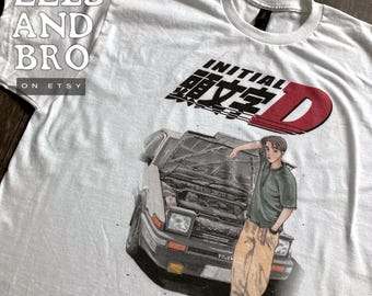 May include: White t-shirt featuring a graphic of a car with its hood open and a person leaning against it. The shirt has the text "INITIAL D" in black and red. The Etsy shop name "EELS AND BRO" is also visible.