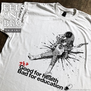 May include: White t-shirt featuring a black and white graphic of two figures in a fight scene, with a gun and blood splatter. Text includes "Good for health, Bad for education" and Japanese characters.