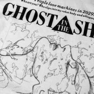 May include: A white t-shirt featuring a black graphic print of the text GHOST IN THE SHELL with a triangle icon. Above the title, the text reads: People love machines in 2029. Who are you? Who slips into my robot body and whispers to my ghost?