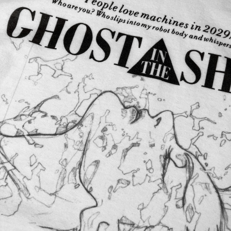 May include: A white t-shirt featuring a black graphic print of the text GHOST IN THE SHELL with a triangle icon. Above the title, the text reads: People love machines in 2029. Who are you? Who slips into my robot body and whispers to my ghost?