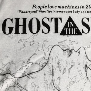 May include: A white t-shirt featuring black text that reads People love machines in 2029 A.D. Who are you? Who slips into my robot body and whispers to me, followed by the title GHOST IN THE SHELL and a black and white line drawing of a figure.