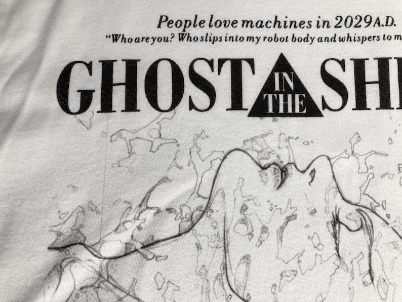 May include: A white t-shirt featuring black text that reads People love machines in 2029 A.D. Who are you? Who slips into my robot body and whispers to me, followed by the title GHOST IN THE SHELL and a black and white line drawing of a figure.