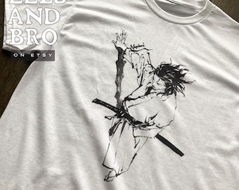 Op de afbeelding: Wit t-shirt met een zwarte inktillustratie van een samoerai met een katana. Het ontwerp bevat het woord "Vagabond" en de tekst "EELS AND BRO ON ETSY" op het shirt.