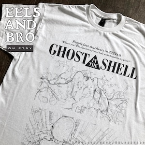 Può includere: T-shirt bianca con la scritta "GHOST IN THE SHELL" in nero, accompagnata da uno schizzo di una persona e di edifici. La maglietta presenta anche la citazione "People love machines in 2029 A.D."