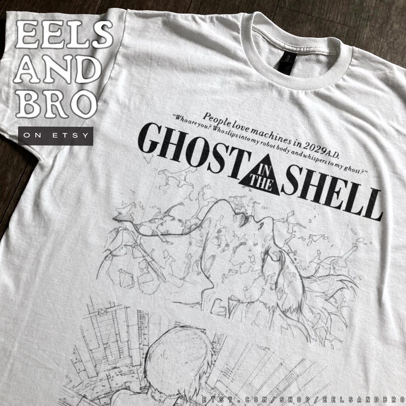 May include: A white short-sleeved t-shirt featuring a black graphic print. The design includes the text "GHOST IN THE SHELL" and the quote "People love machines in 2029 A.D. Who are you? Who slips into my robot body and whispers to my ghost?"