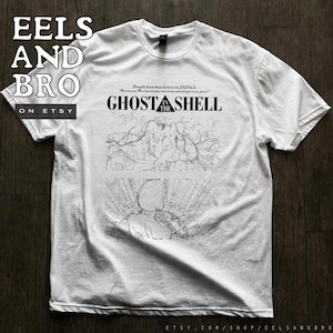 May include: A white short-sleeve t-shirt featuring a black graphic print. The design includes the text GHOST IN THE SHELL, the phrase People love machines in 2029 A.D., and two detailed line art illustrations depicting a figure and a cityscape.